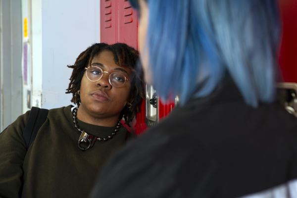 A black genderqueer person and a white person with blue hair talking to each other by some lockers.